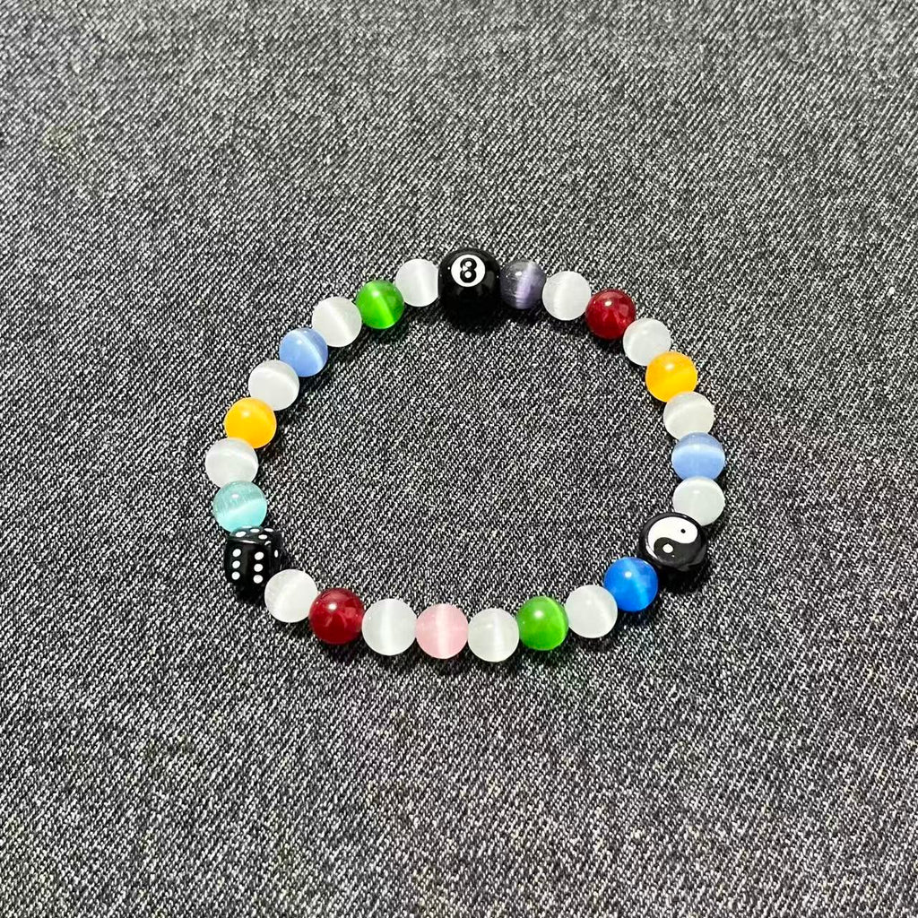 A single SpiritPalette Chroma Rebel bracelet flat lay on dark gray denim fabric, featuring colorful rainbow cat-eye crystal beads with an 8-ball, a yin-yang charm, and a black dice.