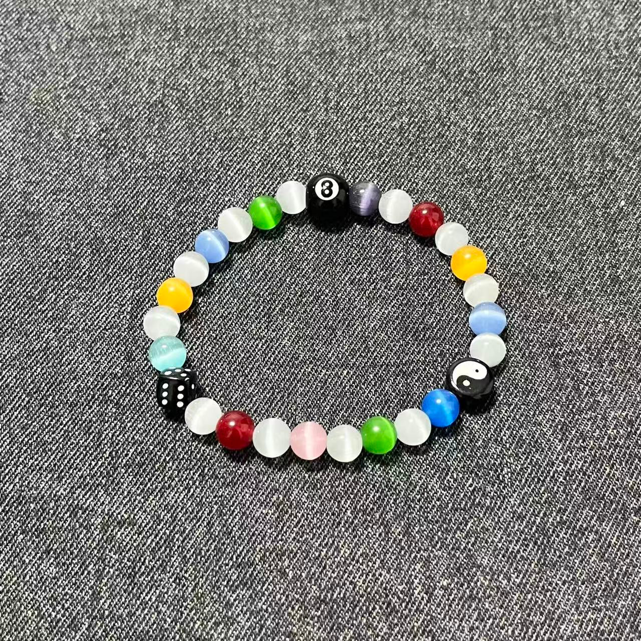 A single SpiritPalette Chroma Rebel bracelet flat lay on dark gray denim fabric, featuring colorful rainbow cat-eye crystal beads with an 8-ball, a yin-yang charm, and a black dice.