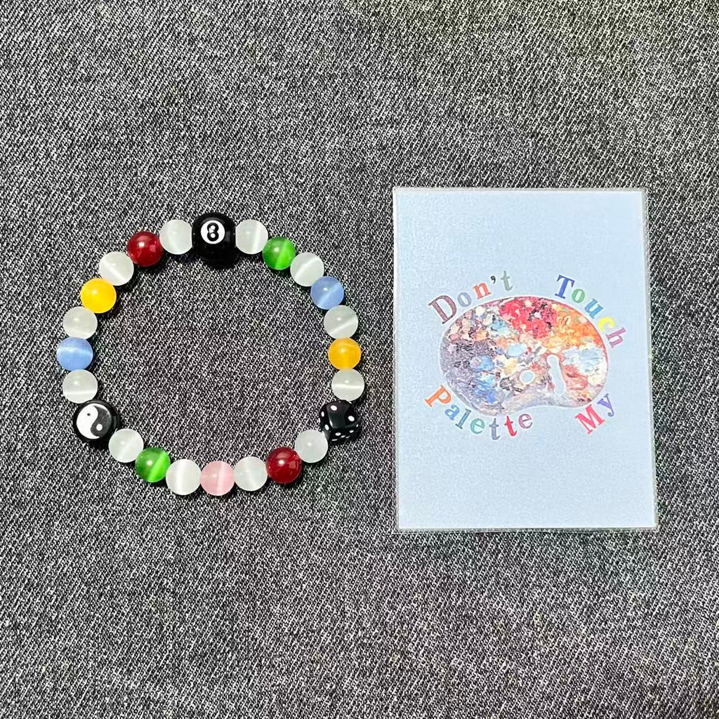 Top-down view of SpiritPalette Chroma Rebel rainbow beaded crystal bracelet next to a Don't Touch My Palette brand art card on gray denim fabric.