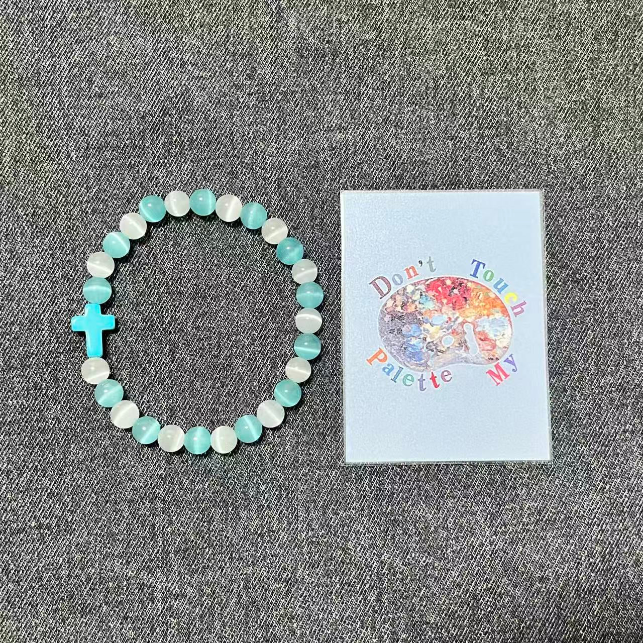 Cyan and white crystal beaded protection bracelet next to SpiritPalette art label on textured gray denim fabric.